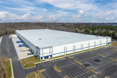 Aerial view of a modern logistics warehouse with loading docks and on-site parking surrounded by wooded landscape.