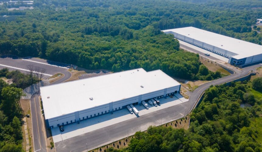 Aerial view of two industrial warehouse buildings surrounded by wooded landscape, with loading docks, truck courts, and parking areas visible.