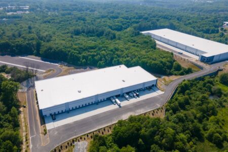 Aerial view of industrial warehouse buildings, showing loading docks, trailer parking, and surrounding wooded landscape.