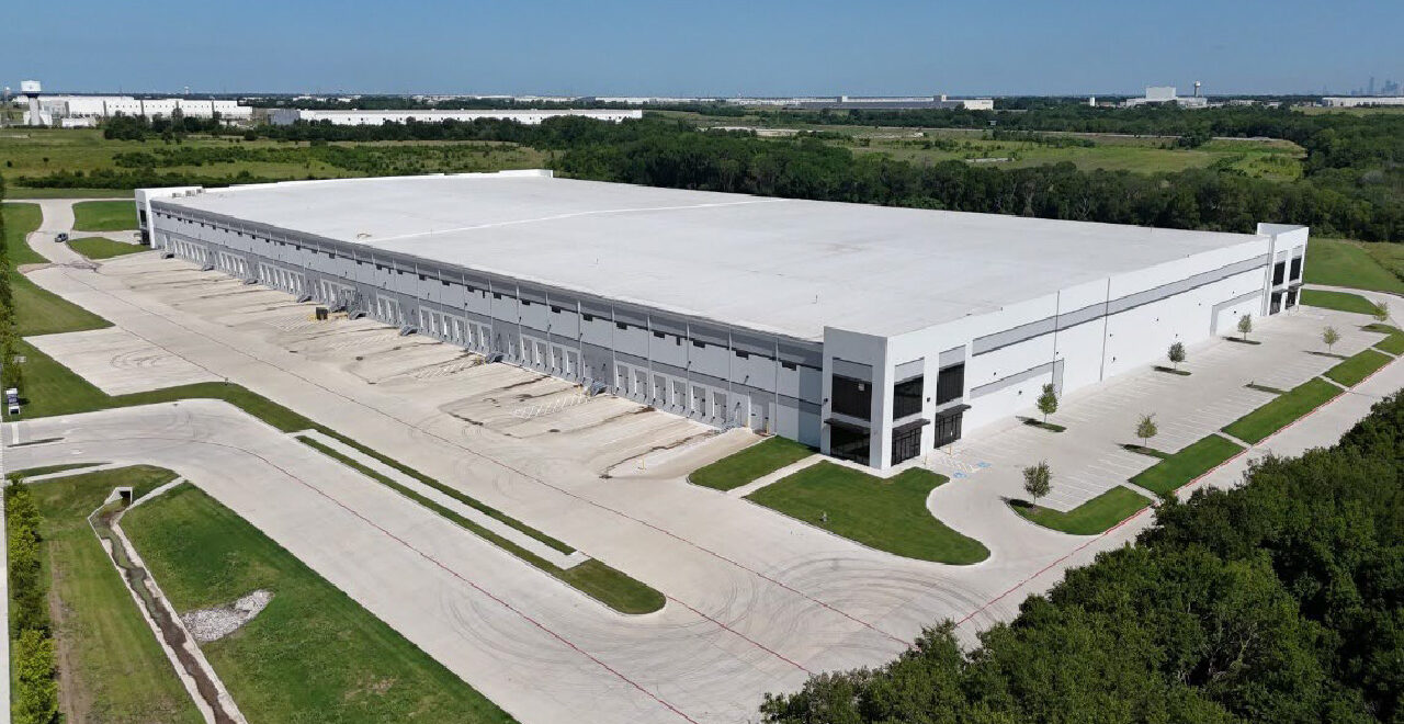 Aerial view of Inland Logistics Center, a large modern white distribution warehouse in Wilmer, Texas, featuring a cross-dock configuration with numerous dock-high loading doors, expansive truck courts, trailer parking areas, and surrounding green space. Industrial buildings and logistics facilities are visible in the background.