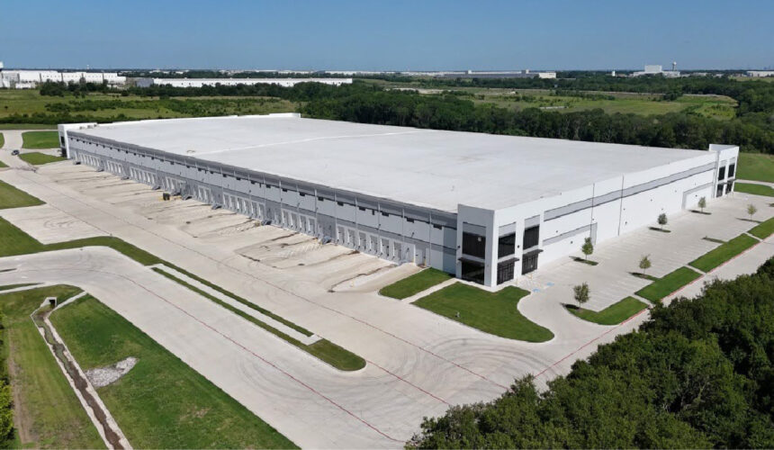 Aerial view of Inland Logistics Center, a large modern white distribution warehouse in Wilmer, Texas, featuring a cross-dock configuration with numerous dock-high loading doors, expansive truck courts, trailer parking areas, and surrounding green space. Industrial buildings and logistics facilities are visible in the background.
